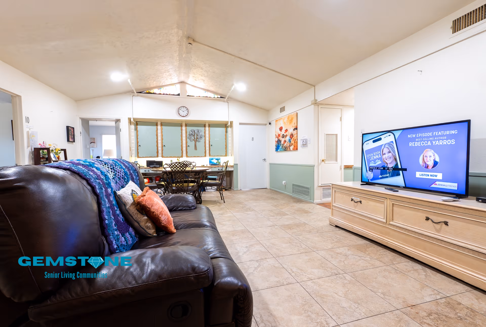 Interior view of a senior living community common area featuring a brown leather couch with colorful pillows and a knitted blanket, a wooden TV stand with a flat-screen TV displaying a podcast episode featuring Rebecca Yarros, a dining table with chairs in the background, and tiled flooring.