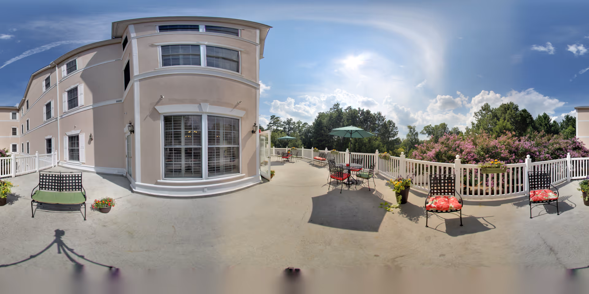 Outdoor patio area at HeartFields at Cary with metal chairs featuring red and green cushions, round tables with green umbrellas, potted plants, white fencing, and a view of trees and flowering bushes under a partly cloudy sky.