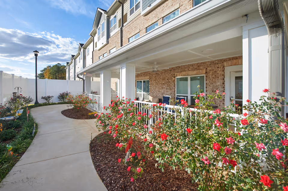 Covered ground-floor porch of a multi-story building with a paved walkway and flowering bushes.