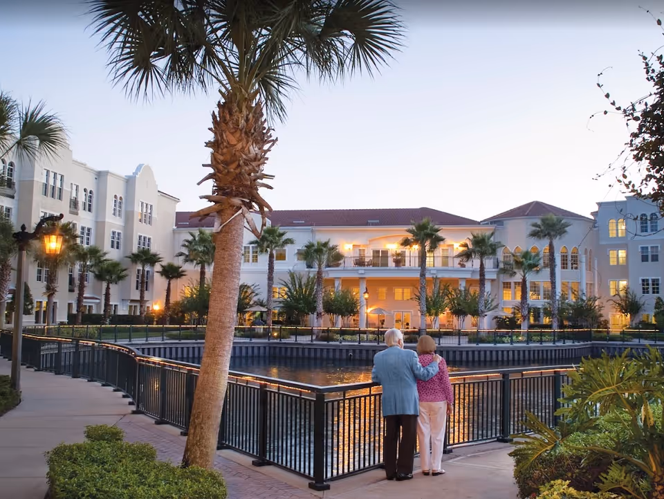 Two people stand by a railing overlooking a pond with palm trees in front of a multi-story senior living building at dusk.