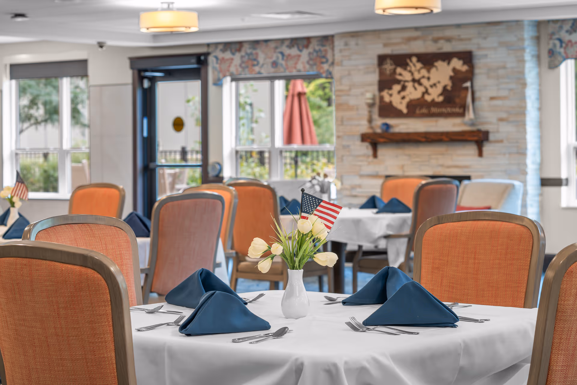 Dining room with round tables set with white tablecloths, blue napkins, a vase of tulips with a small American flag, and orange upholstered chairs.