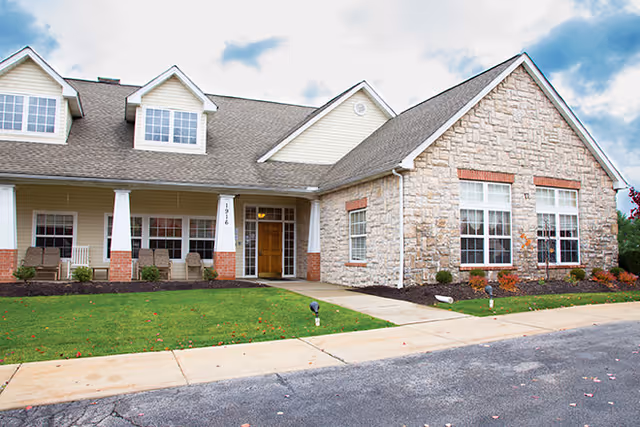 Front exterior of a stone-and-siding senior living building with a covered porch, columns, lawn, and walkway.