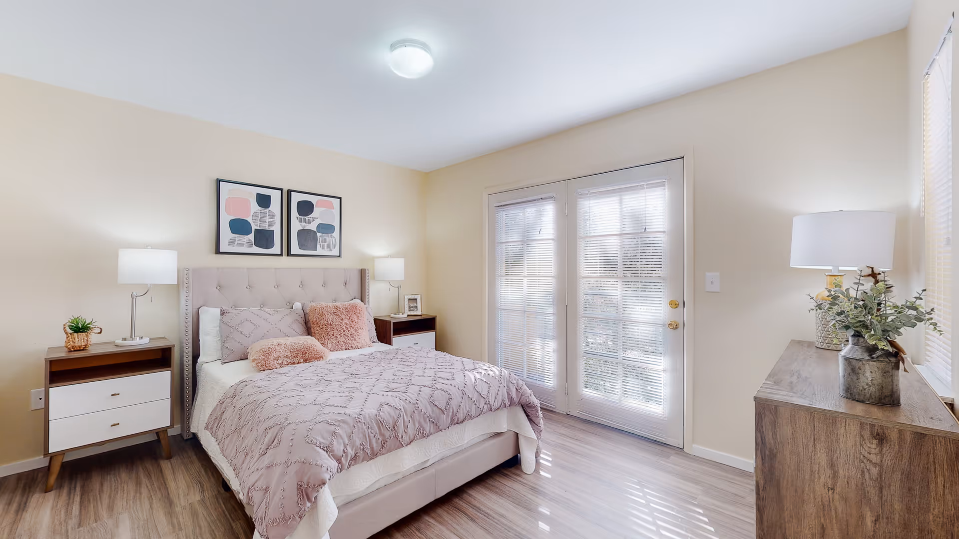 A bright bedroom with a bed featuring a tufted headboard, pink and white bedding, and two pink decorative pillows. There are two nightstands with lamps on either side of the bed, a wooden dresser with a lamp and plant on the right, and French doors with blinds letting in natural light.