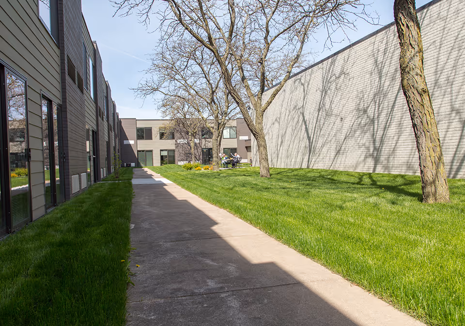 Outdoor walkway between two buildings with green grass and leafless trees on a sunny day. A few people are sitting at a table in the distance near the building.