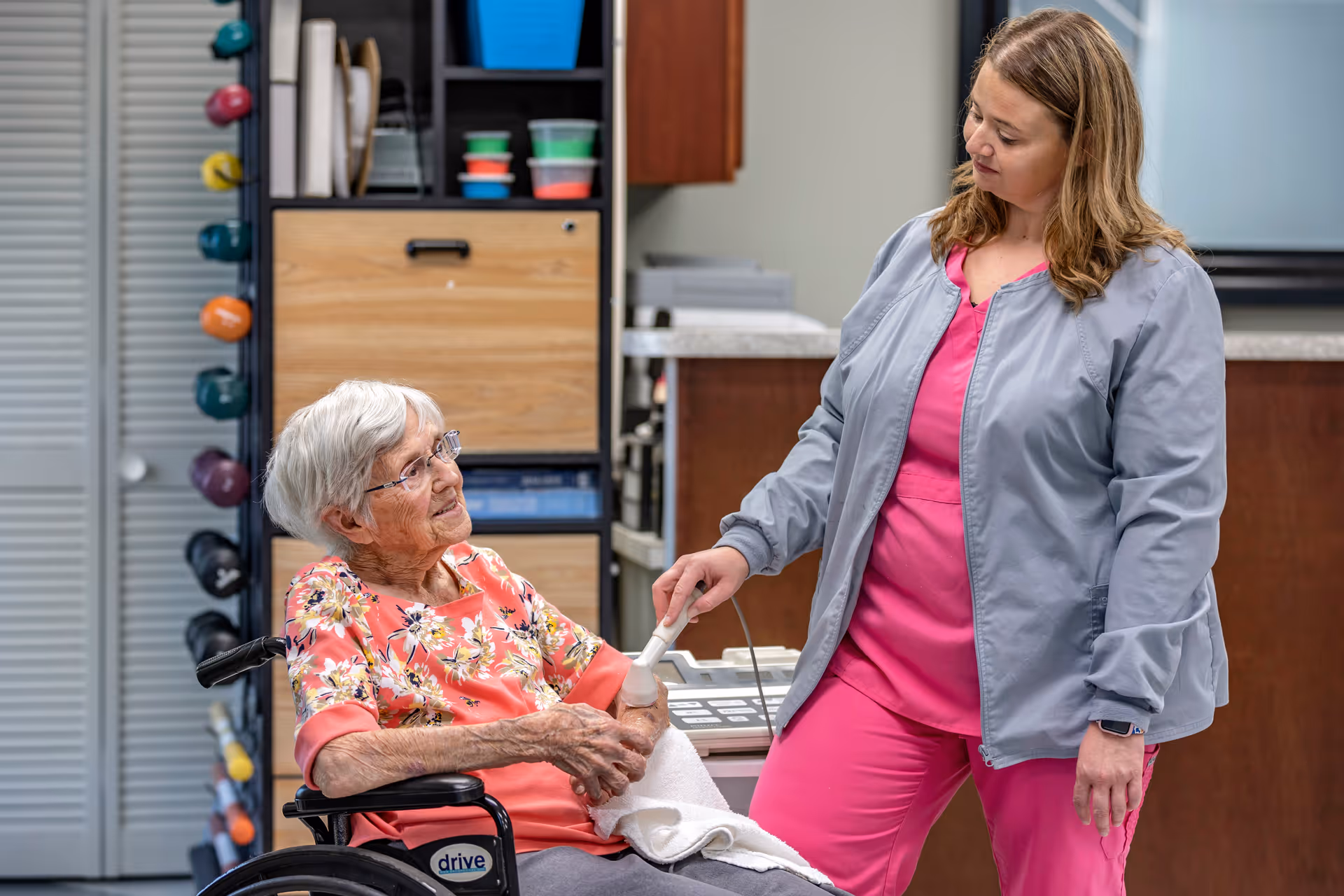 An elderly woman in a wheelchair wearing a floral top is receiving care from a healthcare worker dressed in pink scrubs and a gray jacket, who is holding a medical device to the woman's arm in a clinical setting with shelves and equipment in the background.