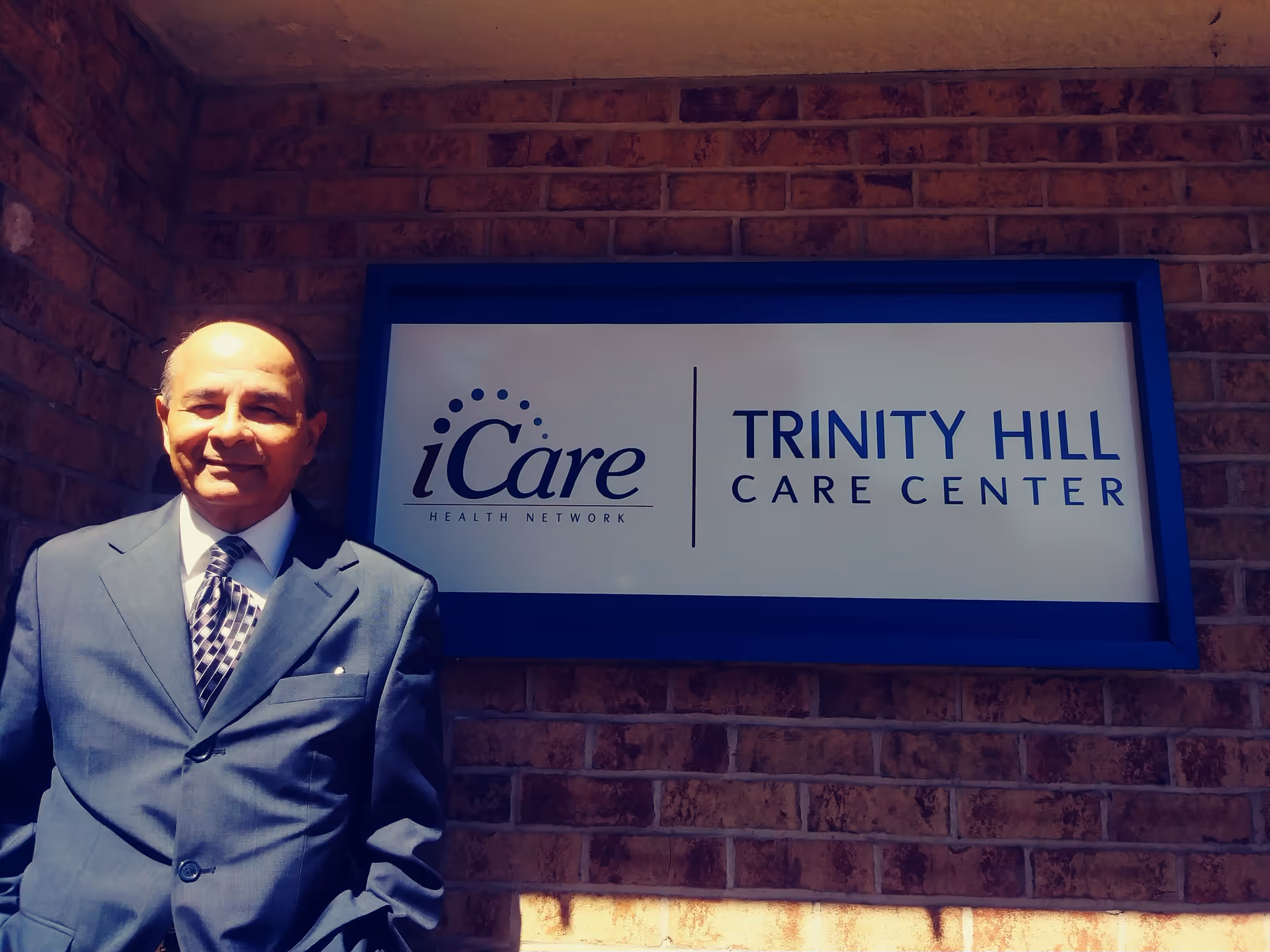 A man in a suit stands next to a 'Trinity Hill Care Center' sign mounted on a brick wall.