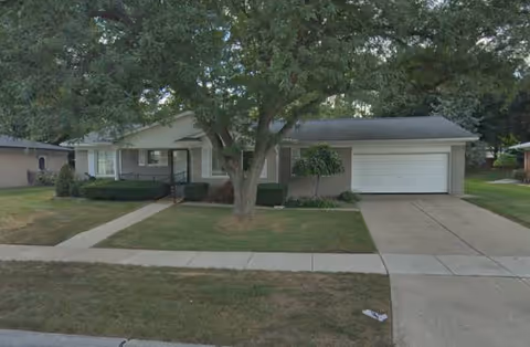 Single-story suburban house with a front yard, driveway, and a large tree.