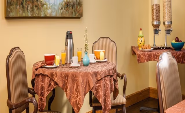 A small round table covered with a patterned brown tablecloth set for breakfast with two chairs. On the table are two cups, two glasses of orange juice, a coffee carafe, and a small vase with a decorative plant. In the background, there is a side table with cereal dispensers, a bowl of fruit, and a decorative ceramic chicken. The walls are painted light yellow and a framed painting hangs above the table.