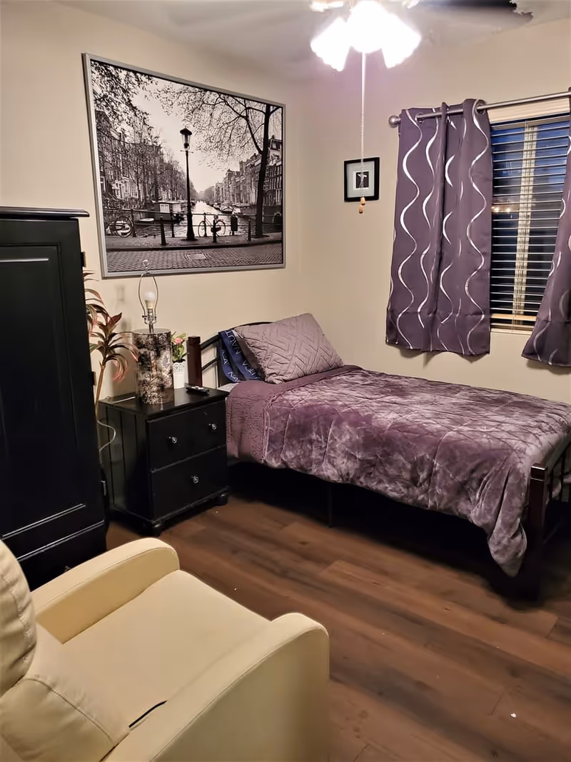 A cozy bedroom with a single bed covered in a purple quilt and matching pillow. Next to the bed is a black nightstand with a lamp and a small plant. A large black and white framed photograph of a canal scene hangs on the wall above the nightstand. Purple curtains with a wavy pattern cover a window with blinds. A beige armchair is partially visible in the foreground. The room has wooden flooring and a ceiling fan with lights.