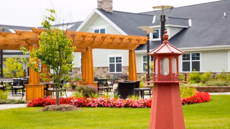 Outdoor garden area at a senior living facility featuring a red lighthouse-style decorative structure in the foreground, green grass, colorful flower beds, a wooden pergola with seating underneath, and a light-colored building with a dark roof in the background.