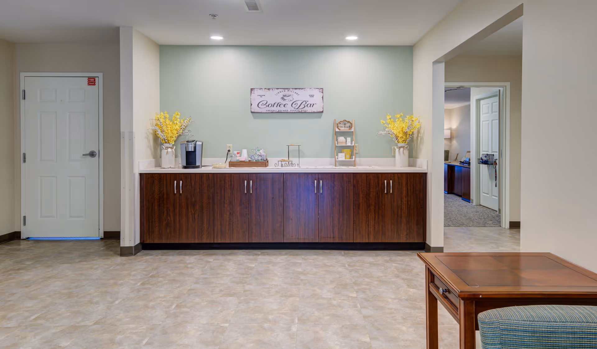 A coffee bar area in a senior living facility with a wooden cabinet, a coffee maker, decorative yellow flowers in vases on both ends, and a sign above that reads 'Coffee Bar'. The room has light-colored walls and flooring, with a doorway leading to another room on the right.
