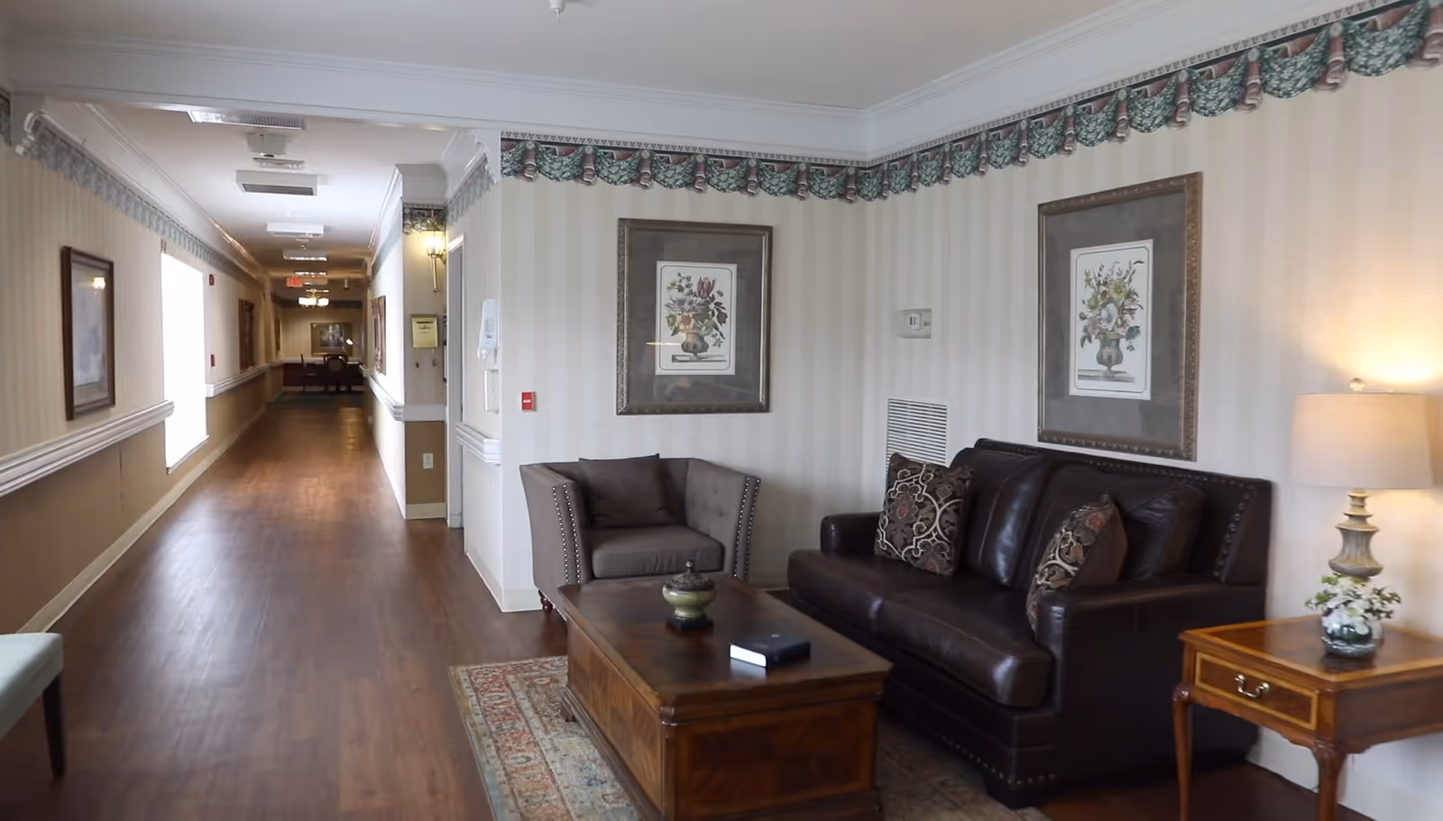 A hallway in a senior living facility with wooden flooring and beige striped wallpaper. On the right side, there is a seating area with a dark brown leather sofa, a gray armchair, a wooden coffee table, and a wooden side table with a lamp and a small plant. Two framed floral paintings hang on the wall above the seating area. The hallway extends into the distance with more framed pictures and lighting fixtures along the walls.