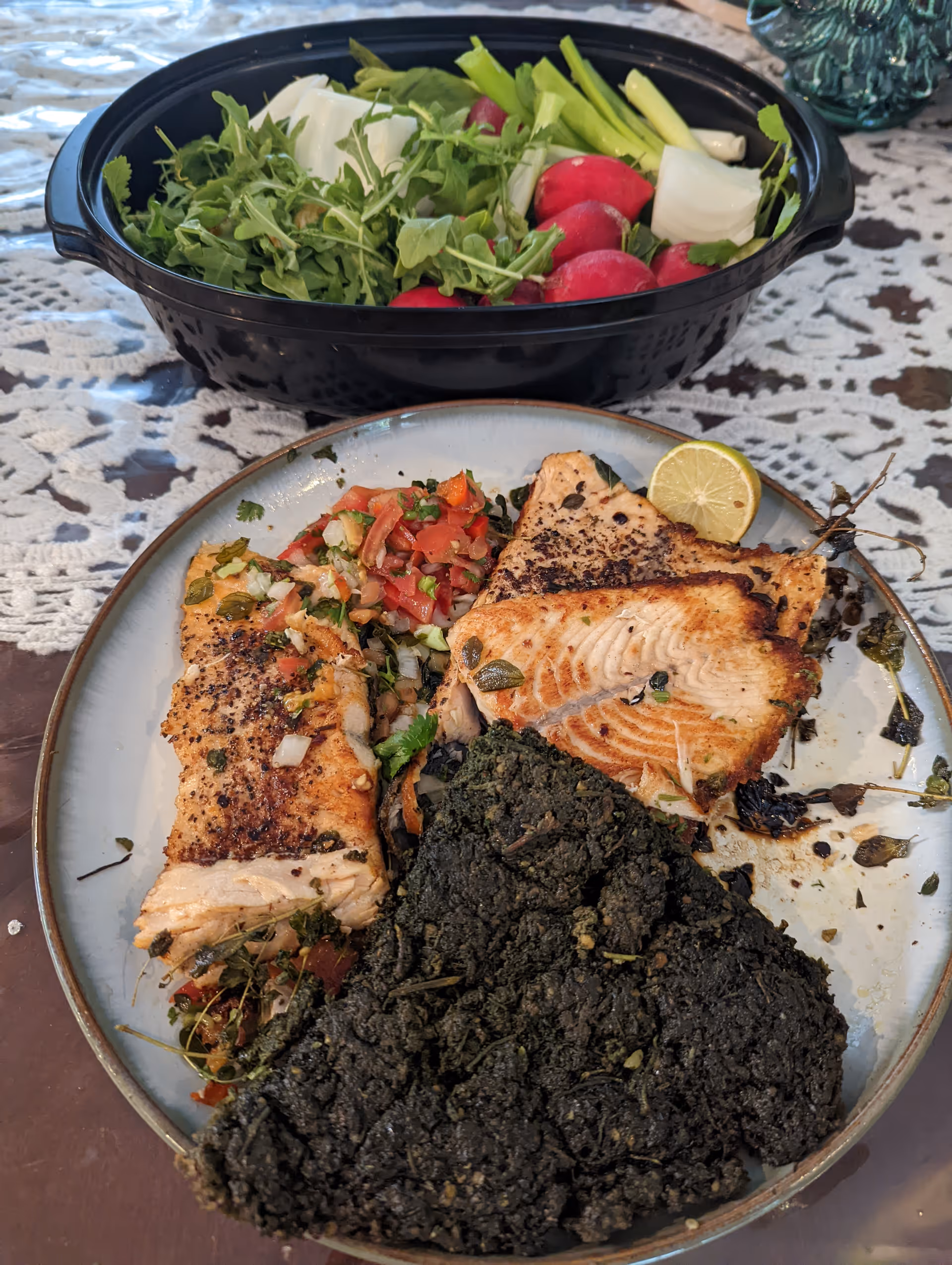 A plate with grilled fish fillets, a serving of cooked leafy greens, and a tomato and onion salsa, accompanied by a black bowl filled with fresh vegetables including radishes, green onions, and leafy greens, all placed on a lace tablecloth.