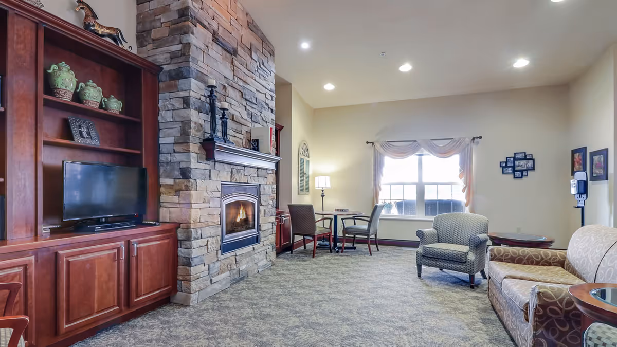 A cozy living room area in Crestwood Village Assisted Living & Memory Care featuring a stone fireplace with a lit fire, a wooden entertainment center with a TV, a small table with two chairs near a window with drapes, an armchair, and a patterned sofa. The room is well-lit with recessed ceiling lights and a floor lamp.