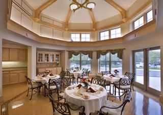 A bright dining room with round tables covered in white tablecloths, each set with plates, napkins, and floral centerpieces. The room features large windows with valances, allowing natural light to fill the space. The ceiling has a decorative wooden beam design with a chandelier hanging in the center. There are wooden cabinets along one wall and glass doors leading outside.