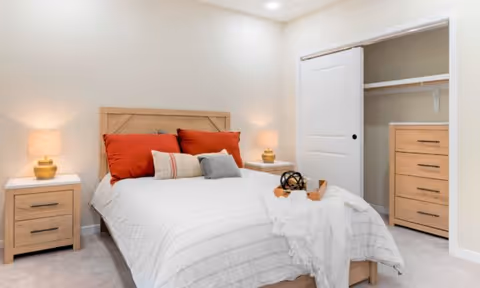A cozy bedroom with a wooden bed frame and white bedding, accented by red and beige pillows. There are two wooden nightstands with matching lamps on either side of the bed. To the right, there is an open closet with sliding doors, showing shelves and drawers inside. The room has soft lighting and a neutral color palette.