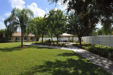 Sunlit outdoor courtyard with a paved path, trees, and a seating area near a light fence.