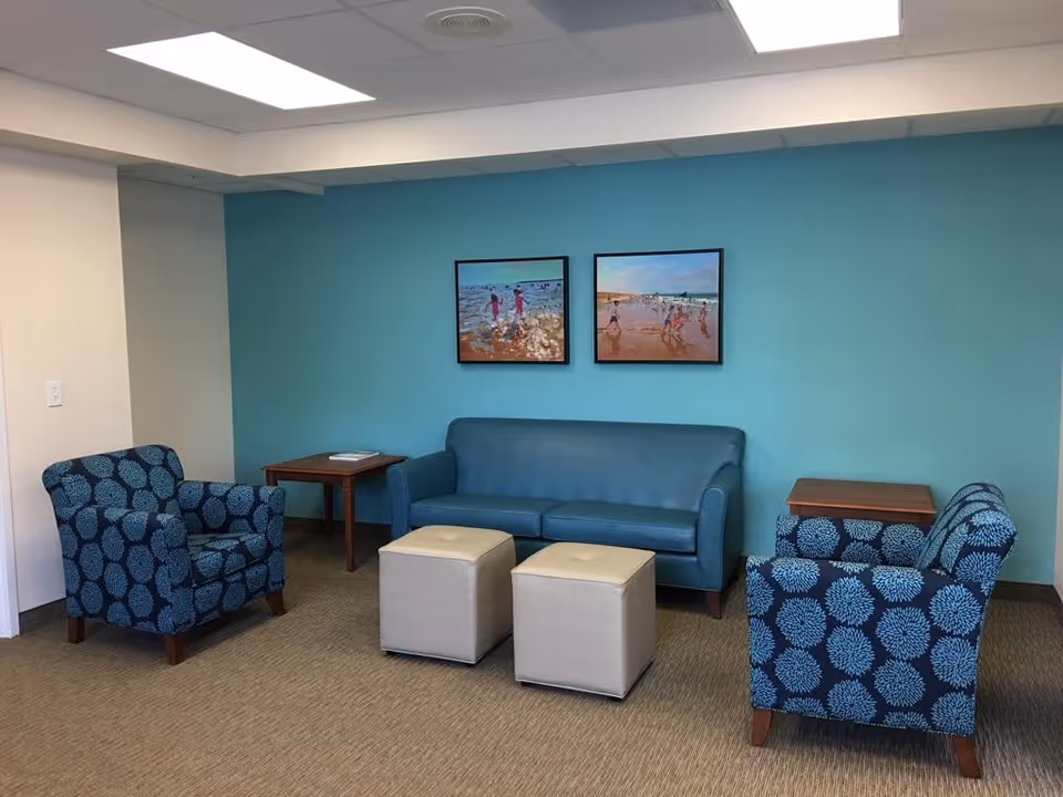 A sitting area with a teal sofa, two blue patterned armchairs, two beige ottomans, and two wooden side tables against a turquoise accent wall with two framed paintings of beach scenes.