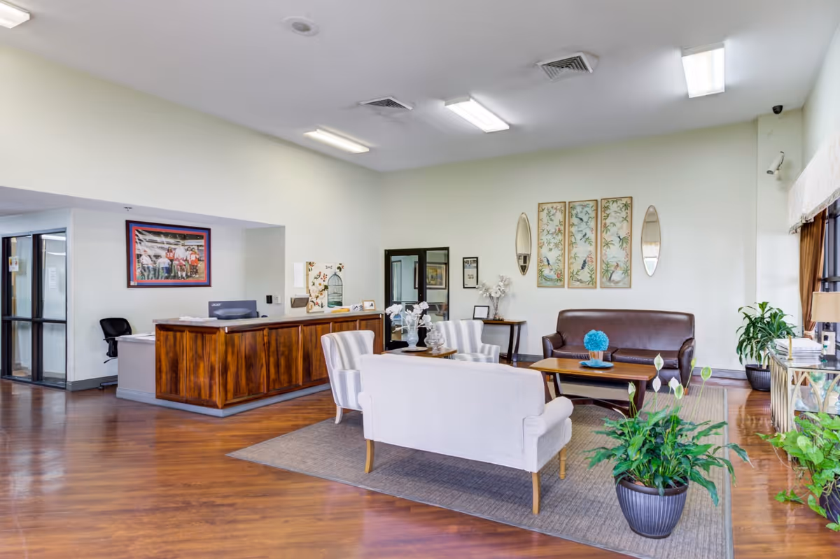 A bright and spacious reception area with a wooden front desk on the left and a seating area on the right. The seating area includes a brown leather sofa, two striped armchairs, and a light-colored loveseat arranged around a wooden coffee table. The room features hardwood floors, several potted plants, framed artwork on the walls, and large windows with curtains allowing natural light to enter.