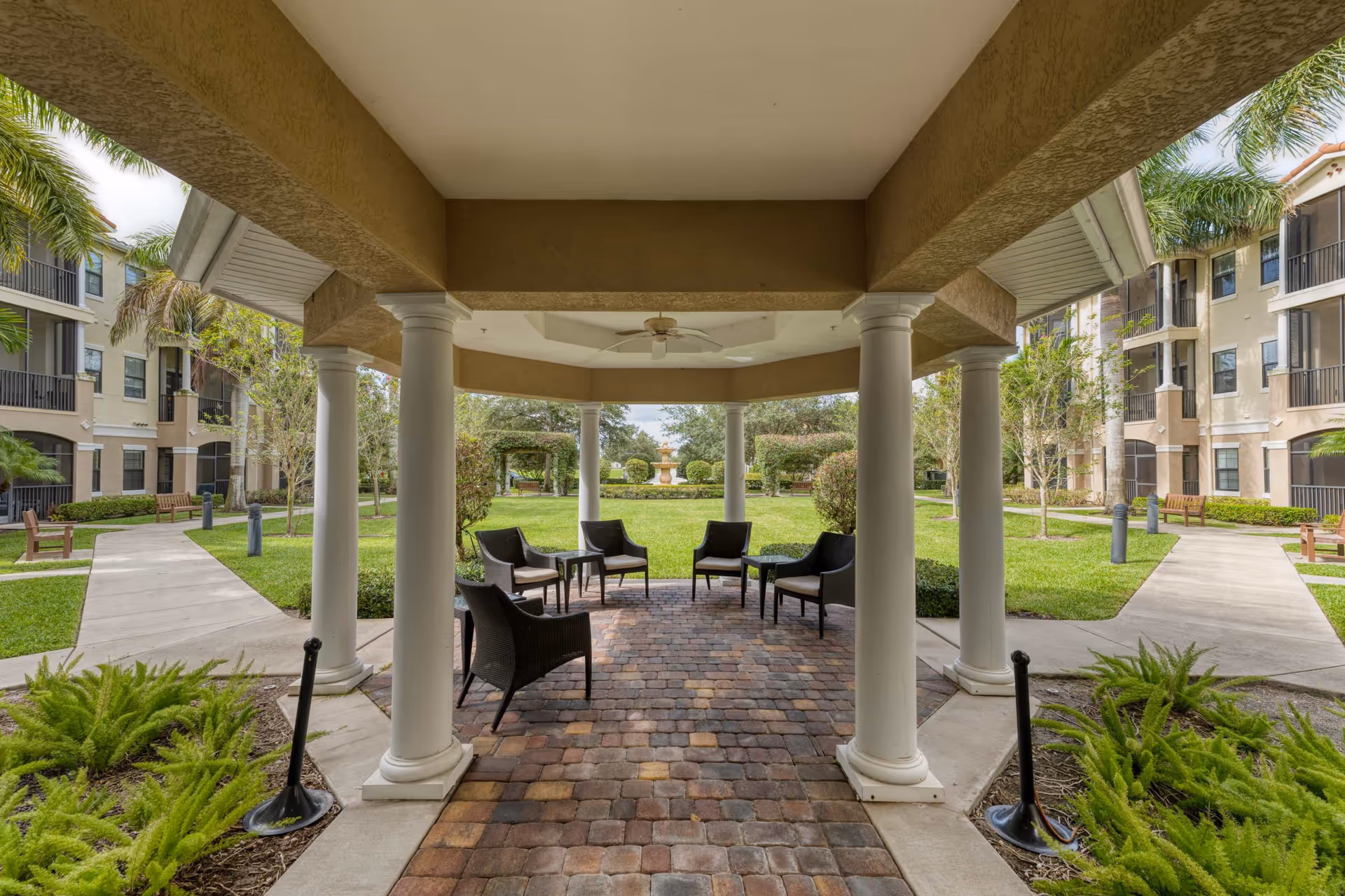 Covered outdoor seating area with white columns and a ceiling fan, surrounded by a paved walkway, green grass, palm trees, and benches. Residential buildings are visible on both sides.