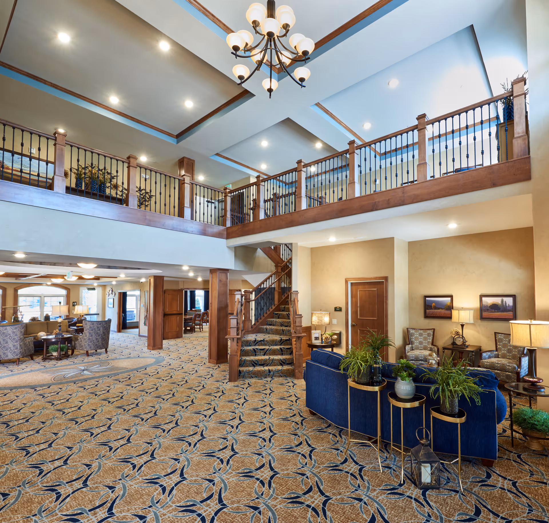Spacious and well-lit senior living facility common area with patterned carpet, a blue sofa with potted plants on side tables, several armchairs, framed artwork on the walls, a wooden staircase leading to an upper balcony with railing, and a chandelier hanging from the ceiling.