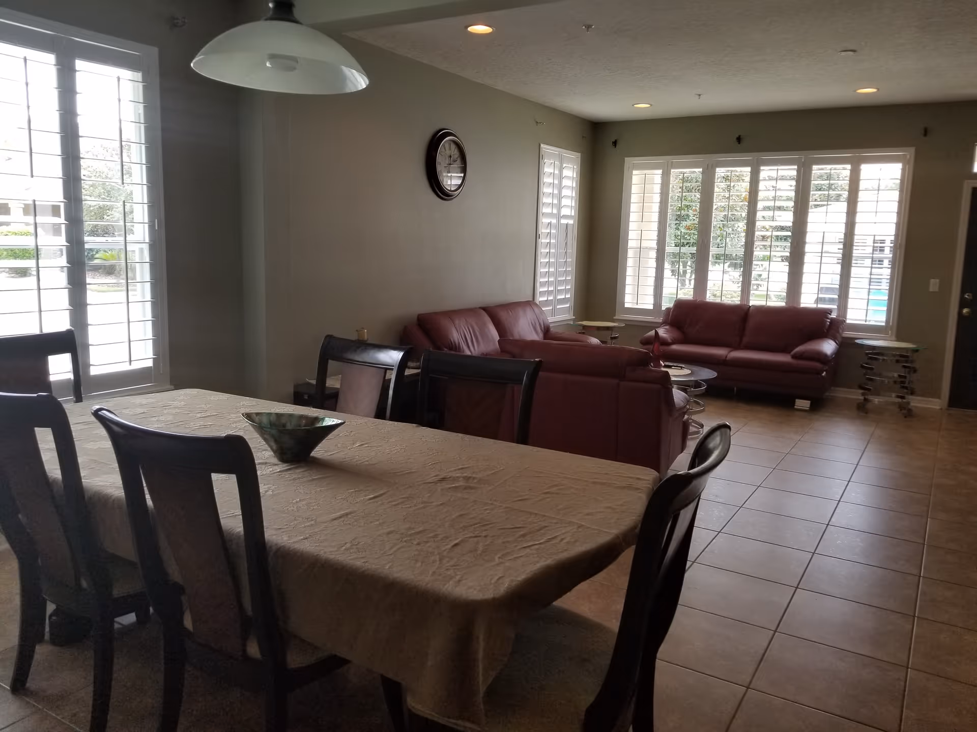 A spacious room with a dining table covered with a beige tablecloth and six chairs around it. In the background, there are three maroon leather sofas arranged near large windows with white shutters. The room has tiled flooring and neutral-colored walls with a clock hanging on one wall. A large white pendant light hangs above the dining table.