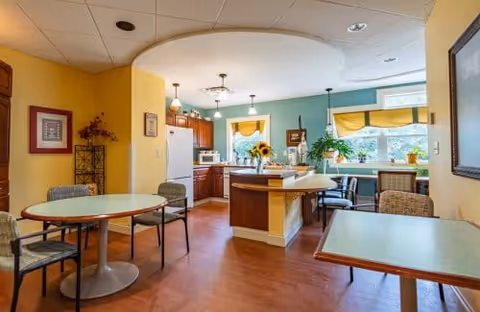 A bright and cozy kitchen and dining area in a senior living facility. The kitchen features wooden cabinets, a white refrigerator, a microwave, and a central island with a vase of sunflowers. The dining area has several tables with chairs, and large windows with yellow valances letting in natural light. The walls are painted in warm yellow and teal colors, creating a welcoming atmosphere.