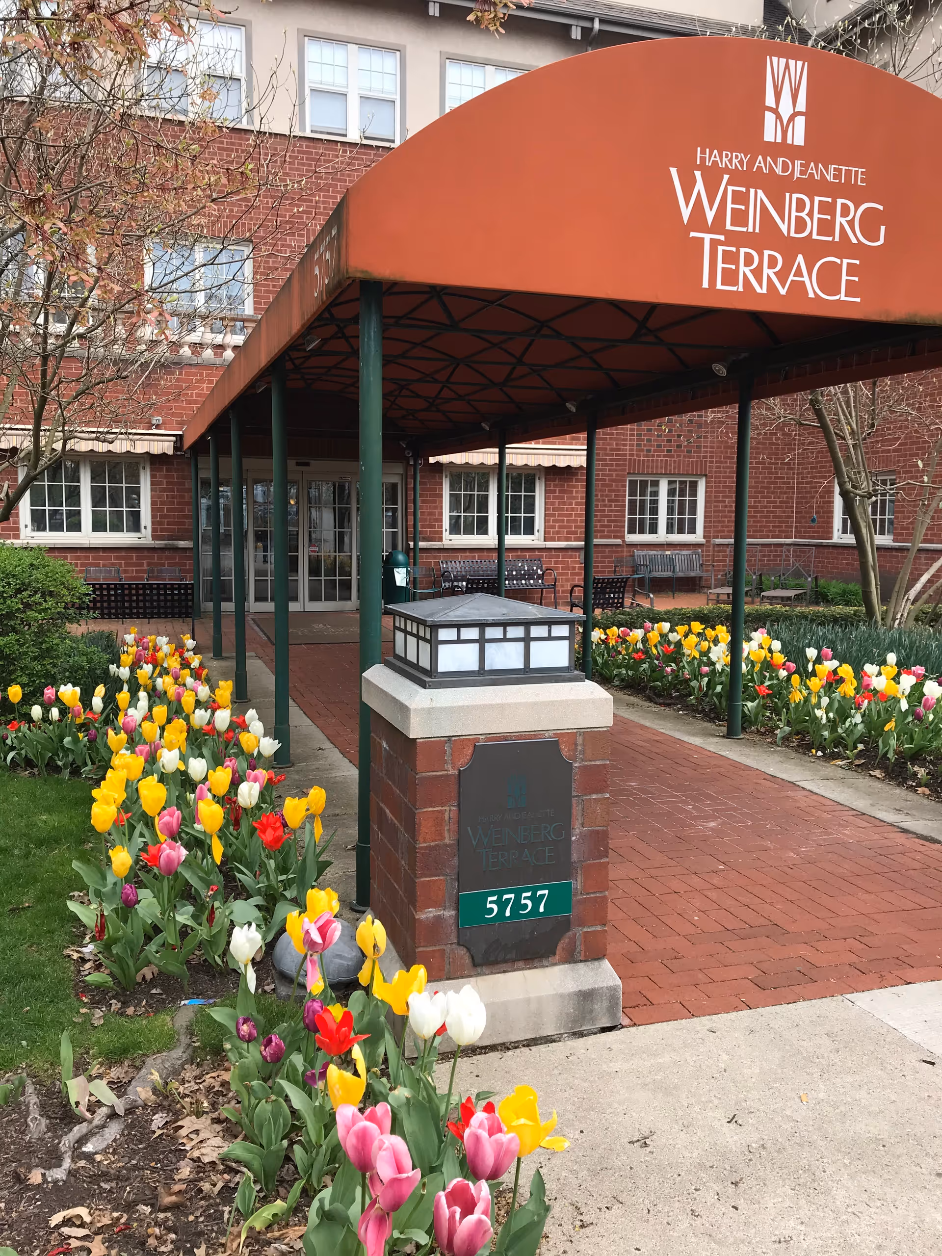 Entrance of Weinberg Terrace with a red awning over a brick walkway, a plaque reading 5757, and colorful tulip flower beds.