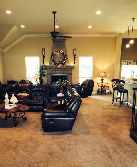 A cozy living room with brown leather sofas arranged around a coffee table. The room features a fireplace decorated with a wreath and string lights, flanked by two windows. There are lamps on side tables and a ceiling fan above. To the right, there is a kitchen counter with bar stools and pendant lights hanging from the ceiling.
