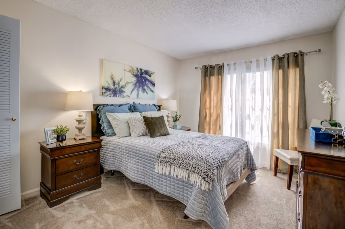 Sunlit bedroom with a made bed topped with decorative pillows, wooden nightstands and lamps, artwork above the headboard, and curtains over a sliding window.