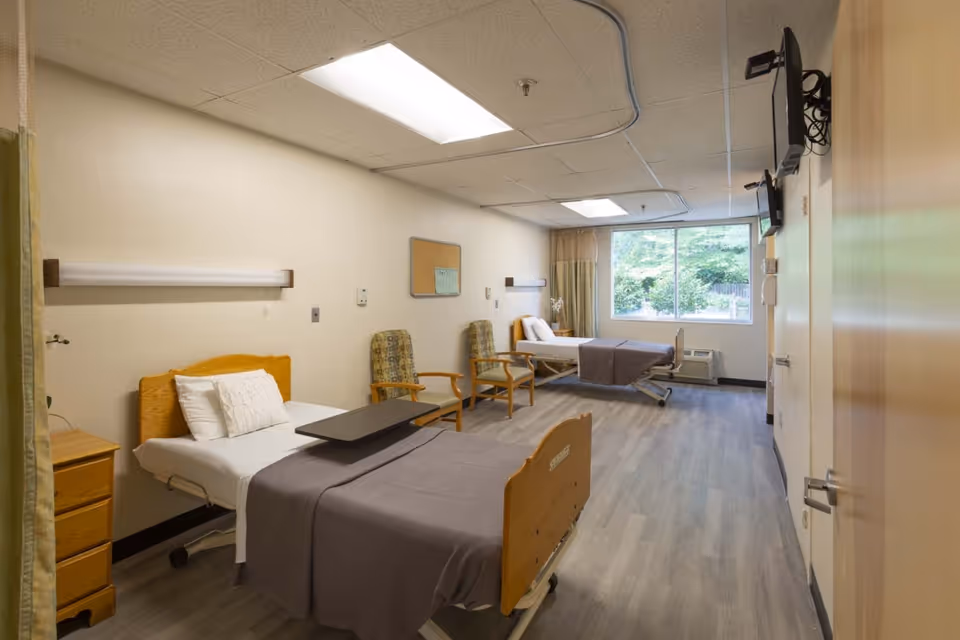 A bright and clean hospital-style room with two beds, each with white pillows and gray blankets. There are two patterned armchairs between the beds, a small wooden nightstand next to the nearest bed, and a large window at the far end letting in natural light. The room has light-colored walls, a wood-patterned floor, and ceiling lights. A television is mounted on the wall near the door.