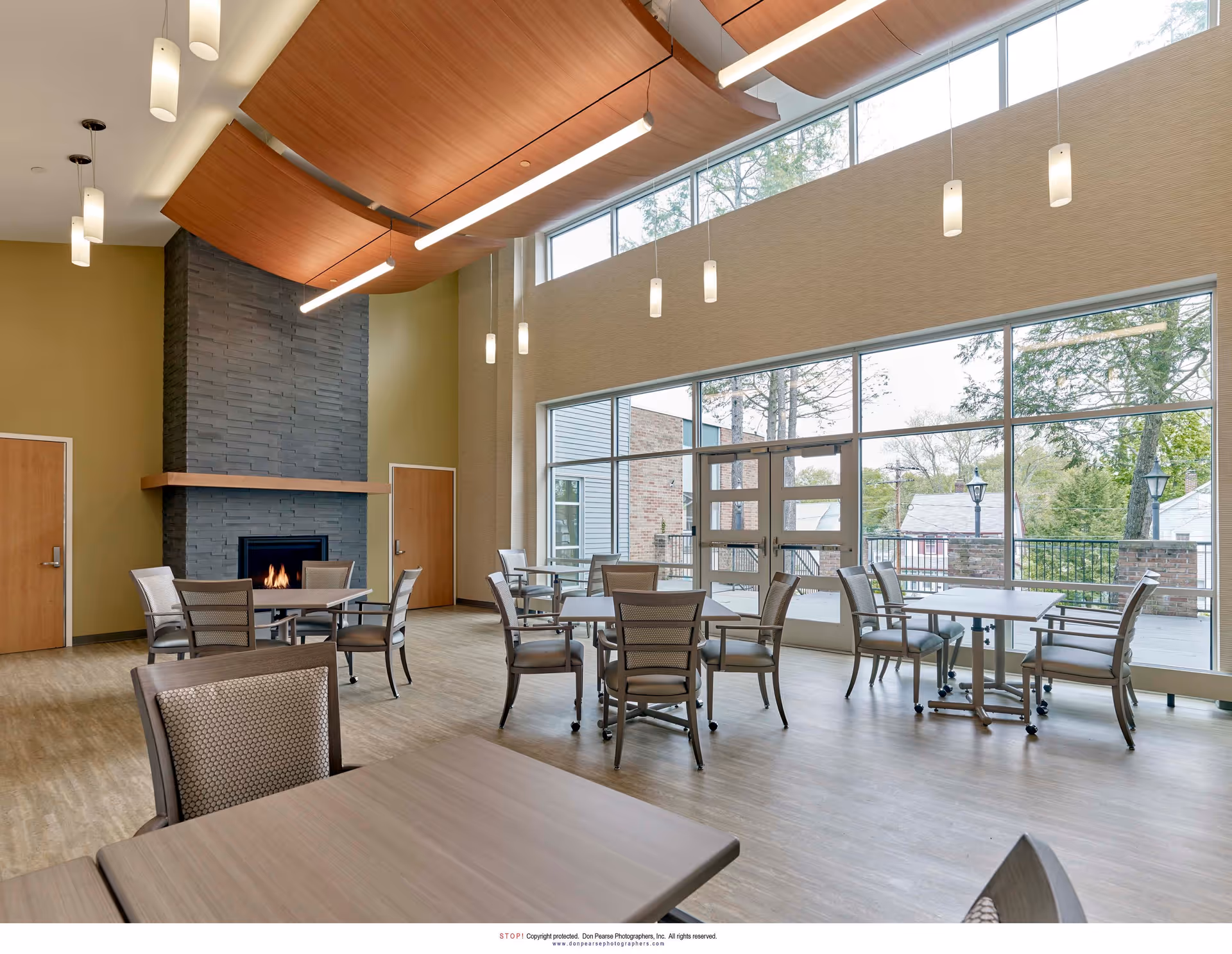 A spacious dining area with multiple tables and chairs arranged neatly. The room features large floor-to-ceiling windows letting in natural light and offering a view of trees and houses outside. There is a modern fireplace with a dark stone surround on one wall, flanked by two wooden doors. The ceiling has unique wooden paneling and hanging pendant lights.