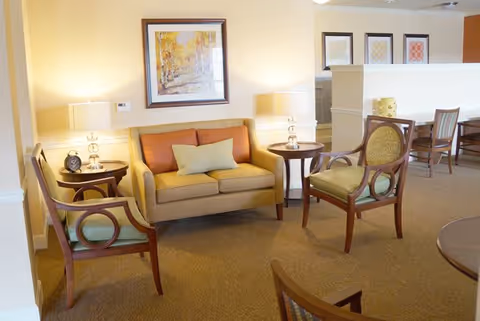 A cozy living room area with a yellow loveseat sofa with a green pillow, two wooden armchairs with green cushions, two round wooden side tables each with a lamp, a framed painting on the wall, and additional seating and artwork visible in the background.