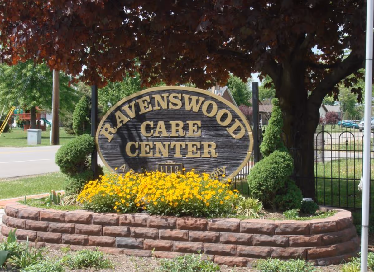 A large wooden sign reading 'Ravenswood Care Center' surrounded by a flower bed with yellow flowers and trimmed bushes, set under a tree with dark red leaves near a street.