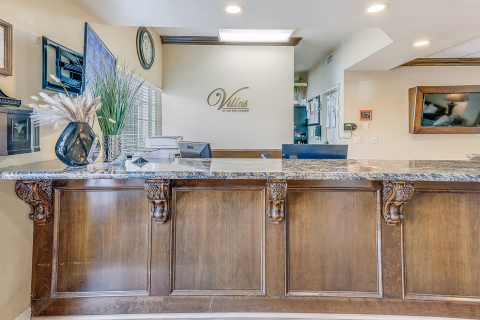 Reception desk area in Villas at San Bernardino facility with a granite countertop, decorative wooden panels, a vase with dried plants, a clock on the wall, and the facility name displayed on the wall behind the desk.