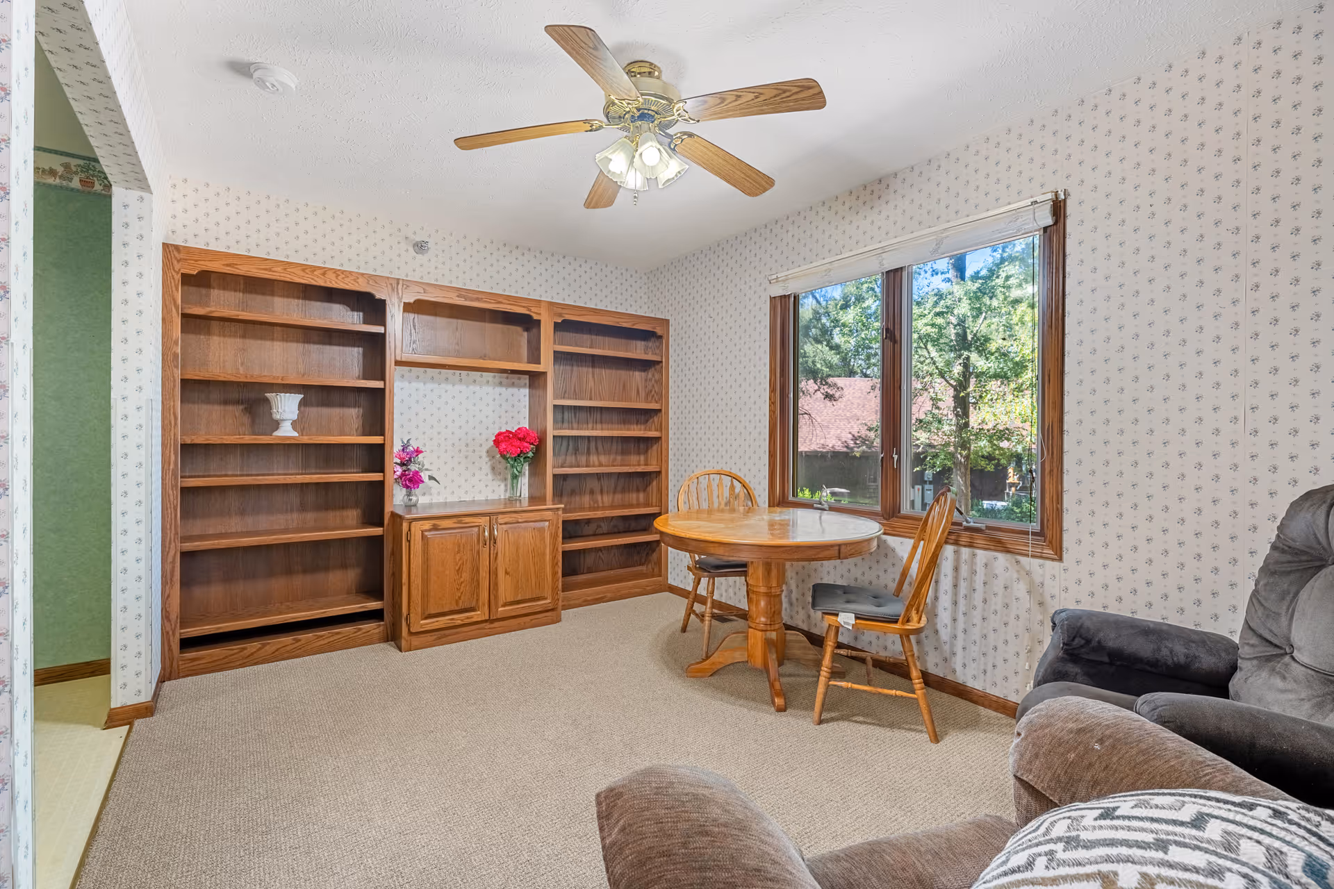 A cozy room with beige carpet and floral wallpaper featuring a wooden round table with two chairs near a large window showing green trees outside. There is a wooden built-in bookshelf with some decorative items and a ceiling fan with lights above. A comfortable armchair is partially visible in the foreground.