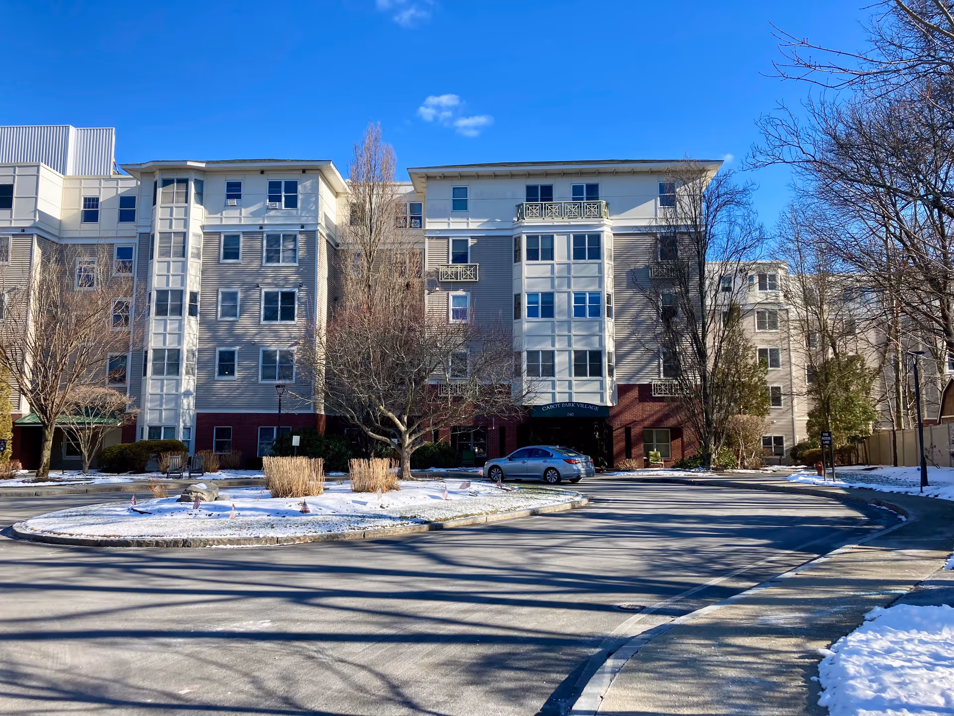 Exterior view of a multi-story senior living facility named Cabot Park Village on a clear winter day with some snow on the ground and leafless trees surrounding the building.