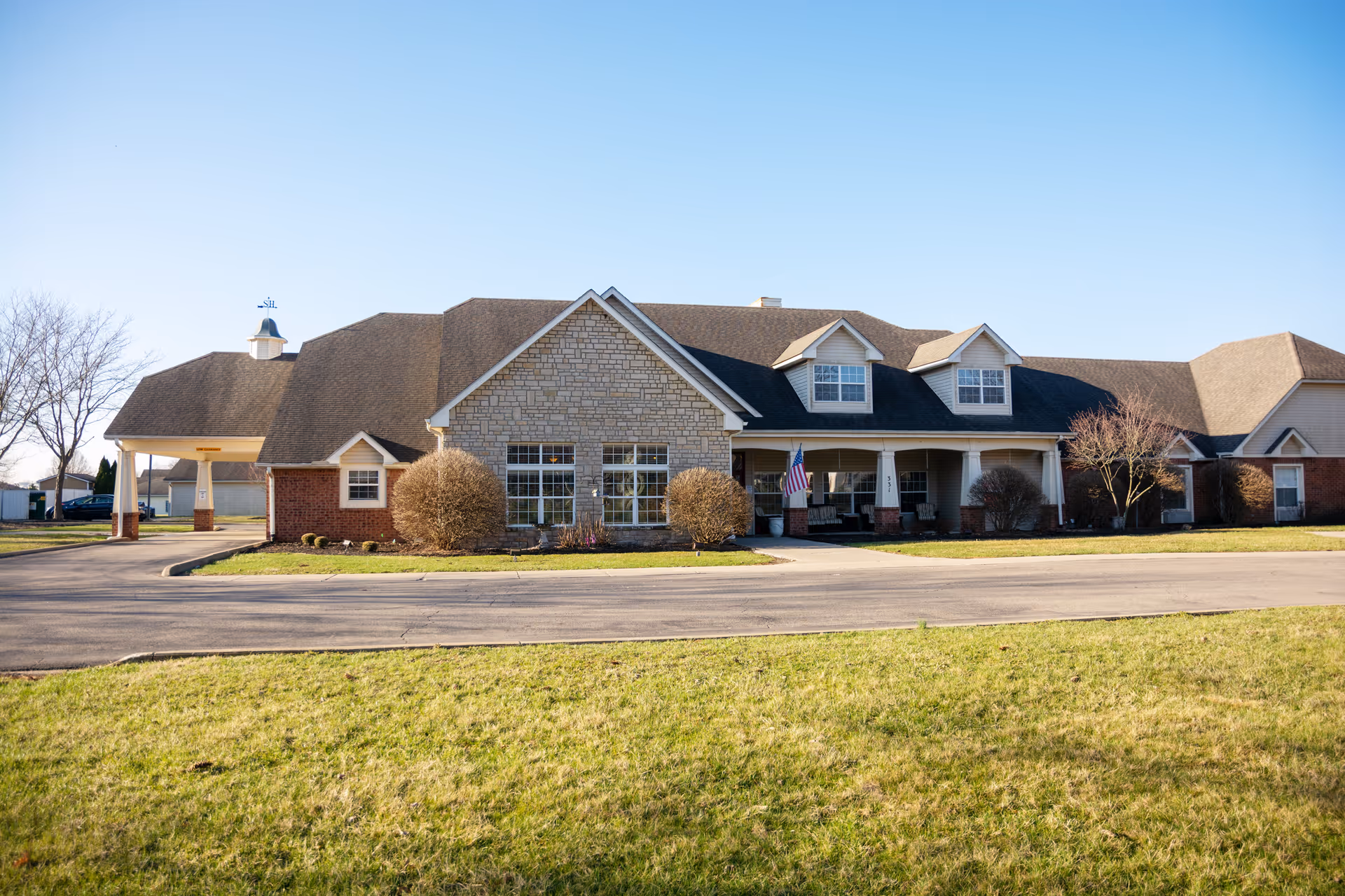 Exterior view of a single-story senior living facility building with a stone and brick facade, multiple windows, a covered entrance with an American flag, and a driveway in front. The building is surrounded by a well-maintained lawn and some leafless trees under a clear blue sky.