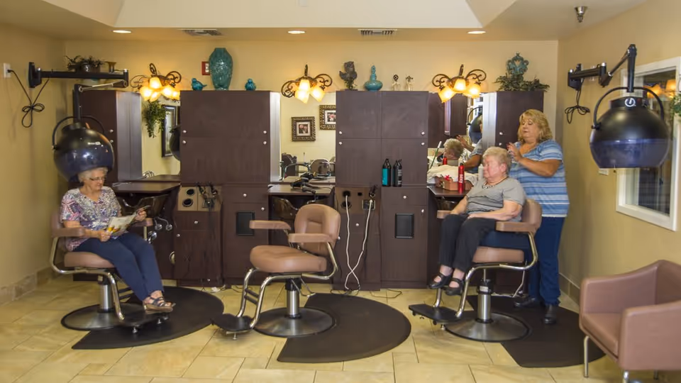 Salon interior with styling stations, hooded hair dryers, two seated older women, and a stylist working on one of them.