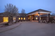 Exterior view of Oak Manor Nursing Center at dusk, showing a single-story brick building with a covered entrance and lit windows. The driveway leads up to the entrance, and there are leafless trees and small landscaped areas around the building.