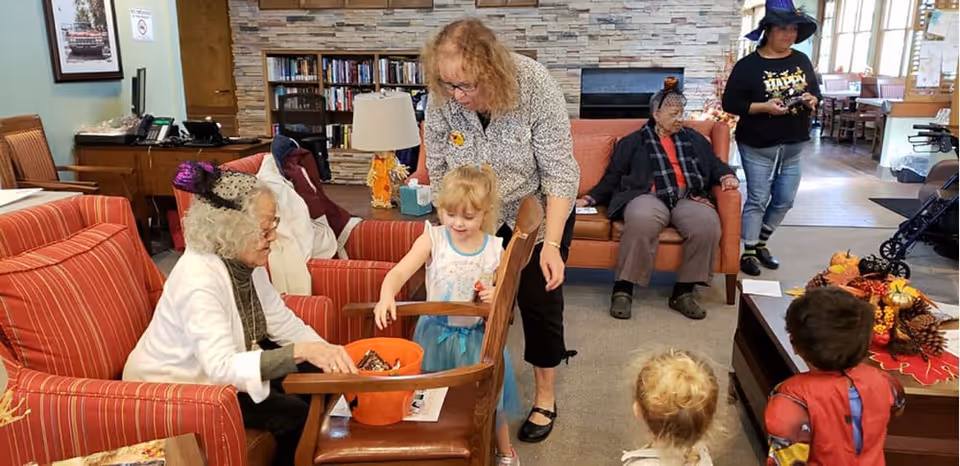 A group of elderly people and children dressed in Halloween costumes are gathered in a cozy living room with red striped armchairs and a stone fireplace. One elderly woman wearing a small purple hat is sitting and handing out candy from an orange bucket to a young girl in a blue dress. Another elderly woman sits on a couch in the background, while a woman stands nearby watching the interaction. Two children in costumes are seen from behind, facing the group. The room has bookshelves, a lamp, and festive autumn decorations on the coffee table.