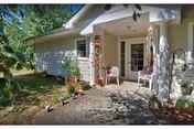 Covered front entrance of a single-story assisted living building with a small porch, chairs, potted plants, and a concrete walkway.