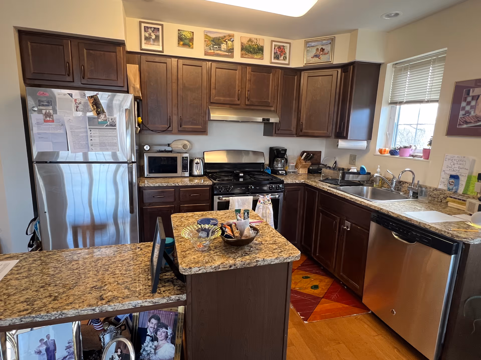 A kitchen with dark wood cabinets, granite countertops, and stainless steel appliances including a refrigerator, stove, microwave, and dishwasher. There is a small island in the center with snacks and a glass bowl on top. Several framed pictures are hung above the cabinets. A window with blinds is above the sink, and various kitchen items are placed on the counters.