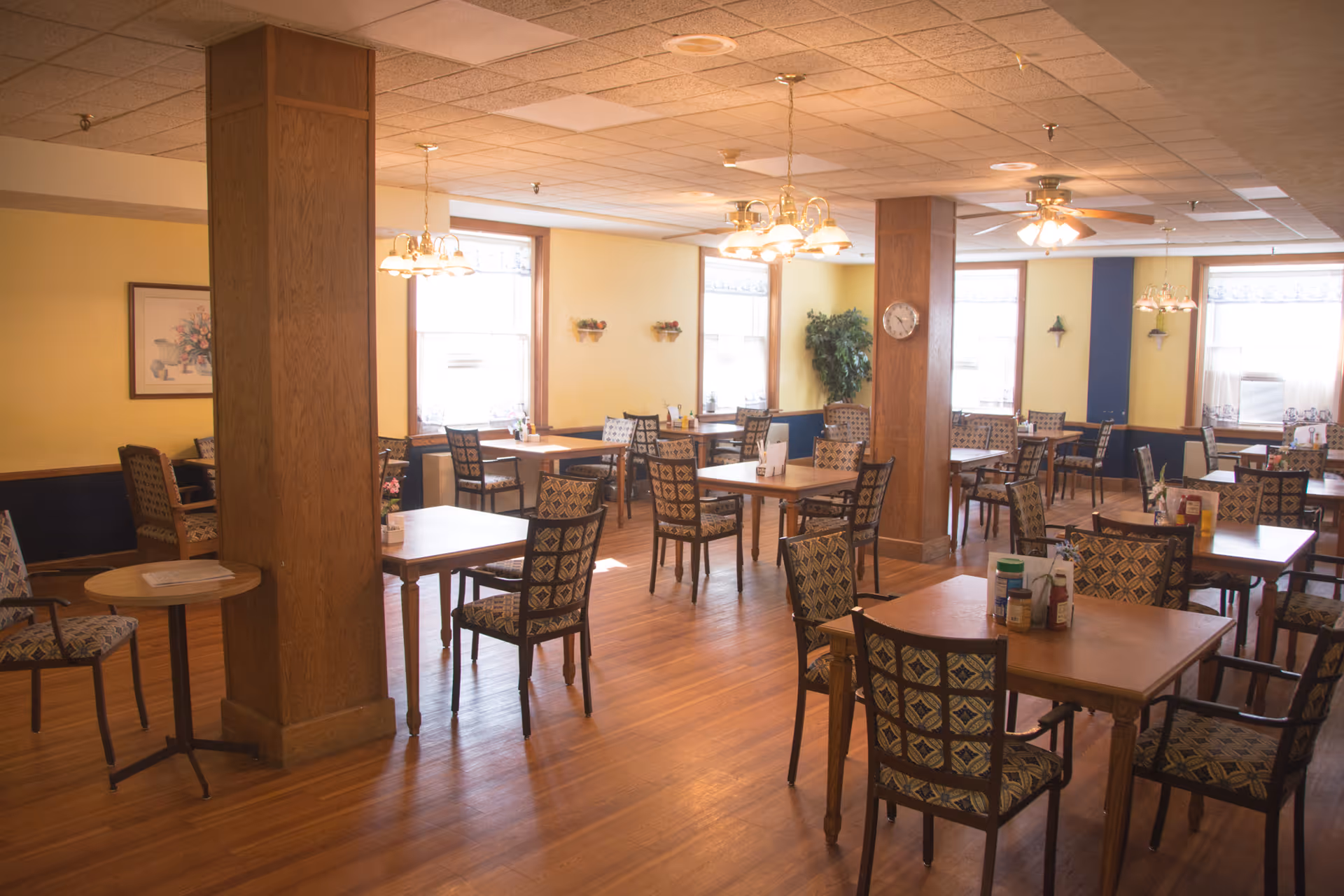 A bright dining room with multiple wooden tables and patterned chairs arranged neatly. The room has large windows letting in natural light, yellow walls with blue wainscoting, wooden pillars, ceiling fans, and hanging light fixtures. There are condiments on the tables and some wall decorations including a clock and framed artwork.