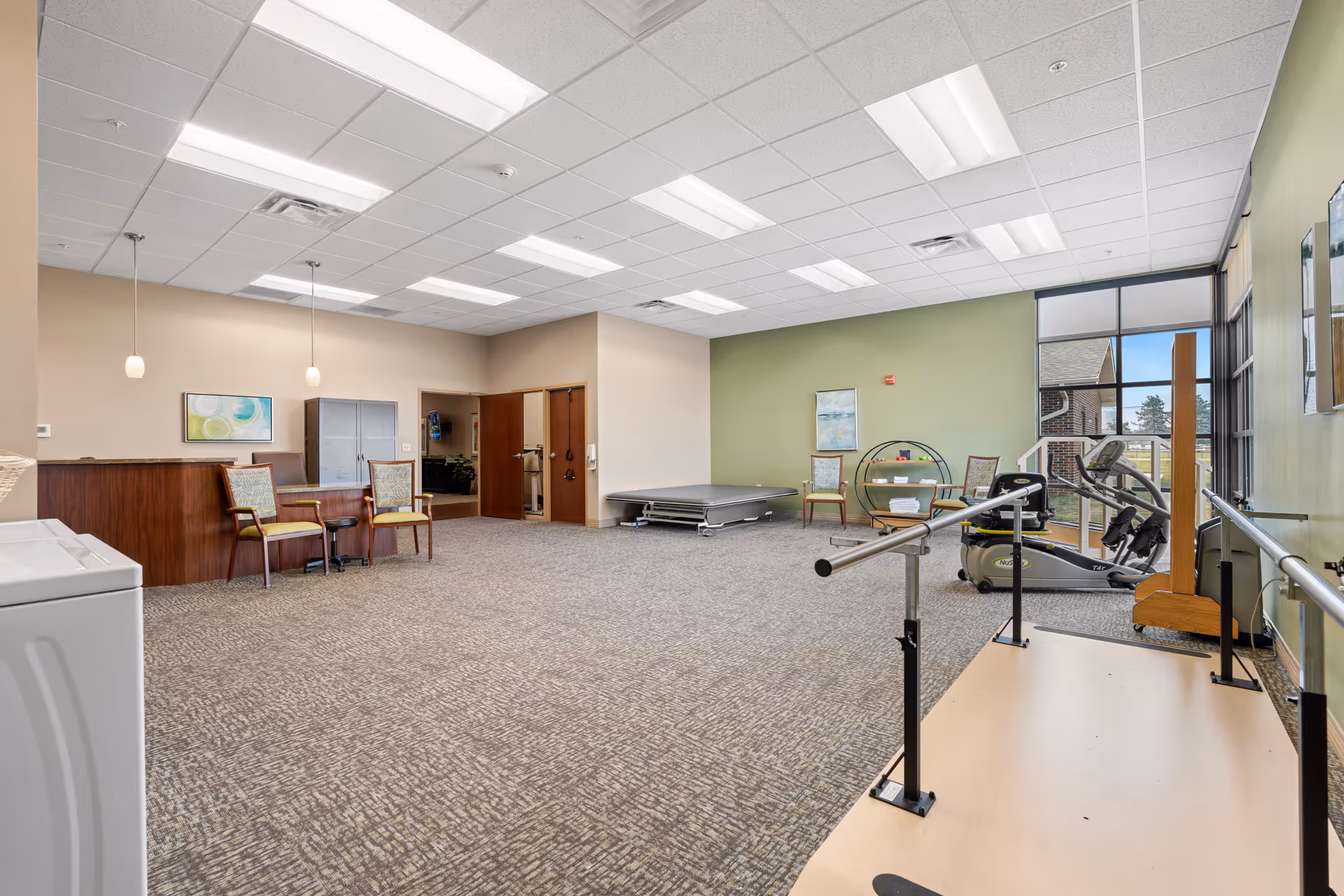 A spacious rehabilitation or physical therapy room with exercise equipment including a stationary bike and parallel bars. The room has carpeted flooring, a green accent wall, several chairs, a small table, and large windows letting in natural light. There are ceiling lights and two pendant lights hanging over a wooden counter area.