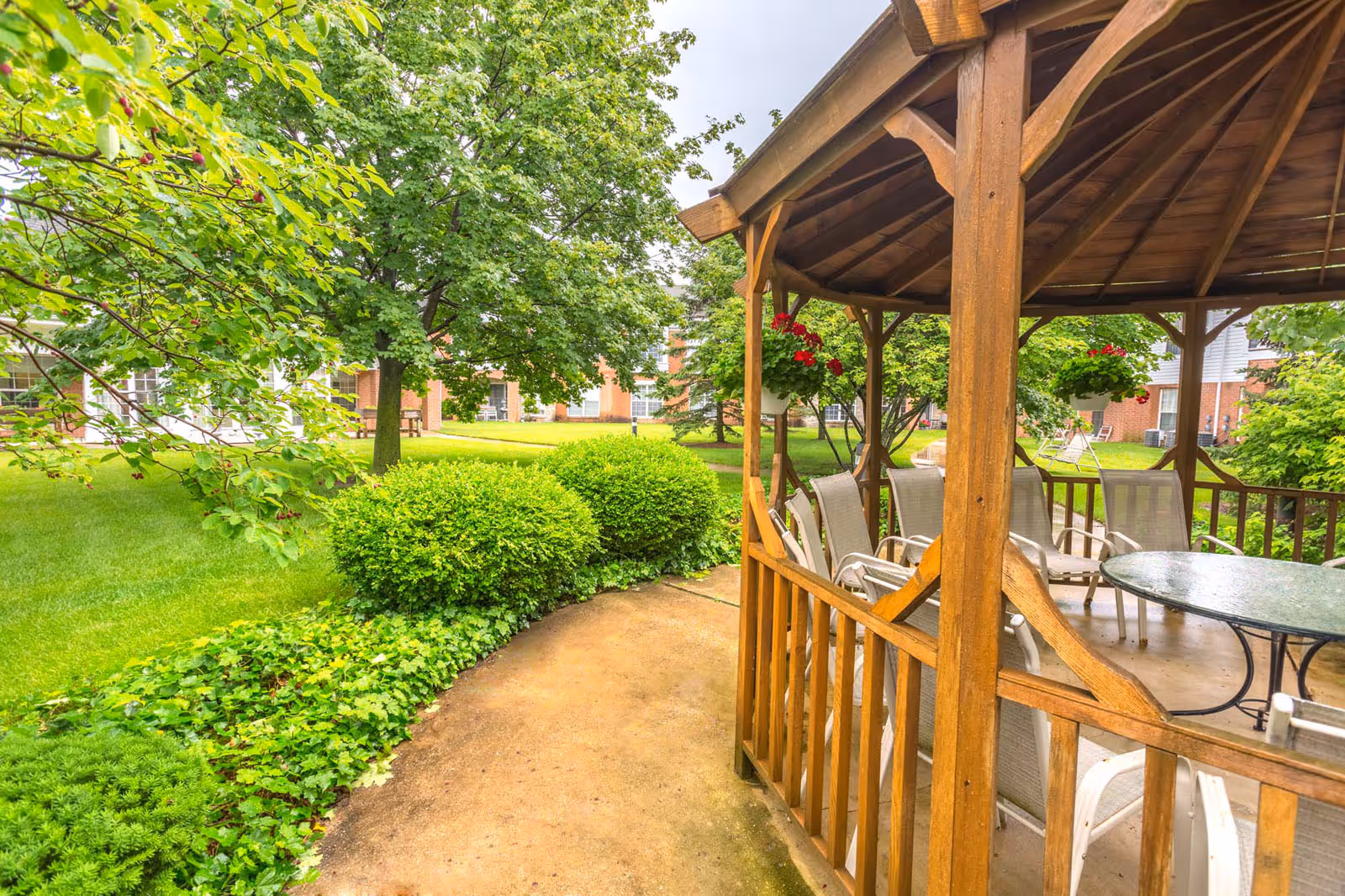Outdoor garden area with a wooden gazebo containing a glass table and several chairs. The surrounding area features green grass, bushes, trees, and hanging flower pots with red flowers. In the background, there are brick buildings with windows and a bench.