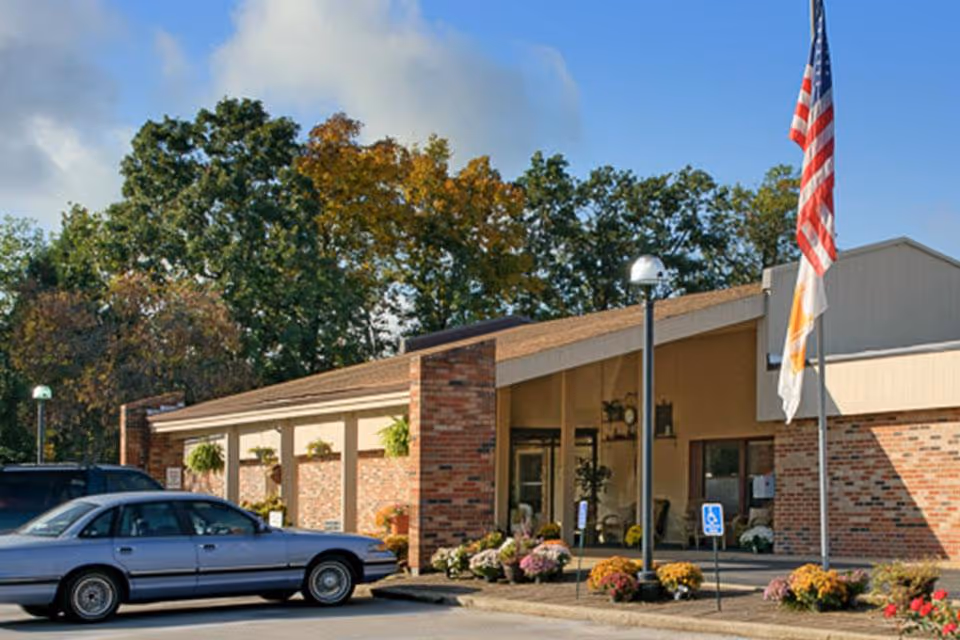 Front entrance of a brick senior living facility with parked cars, flags, and decorative landscaping.