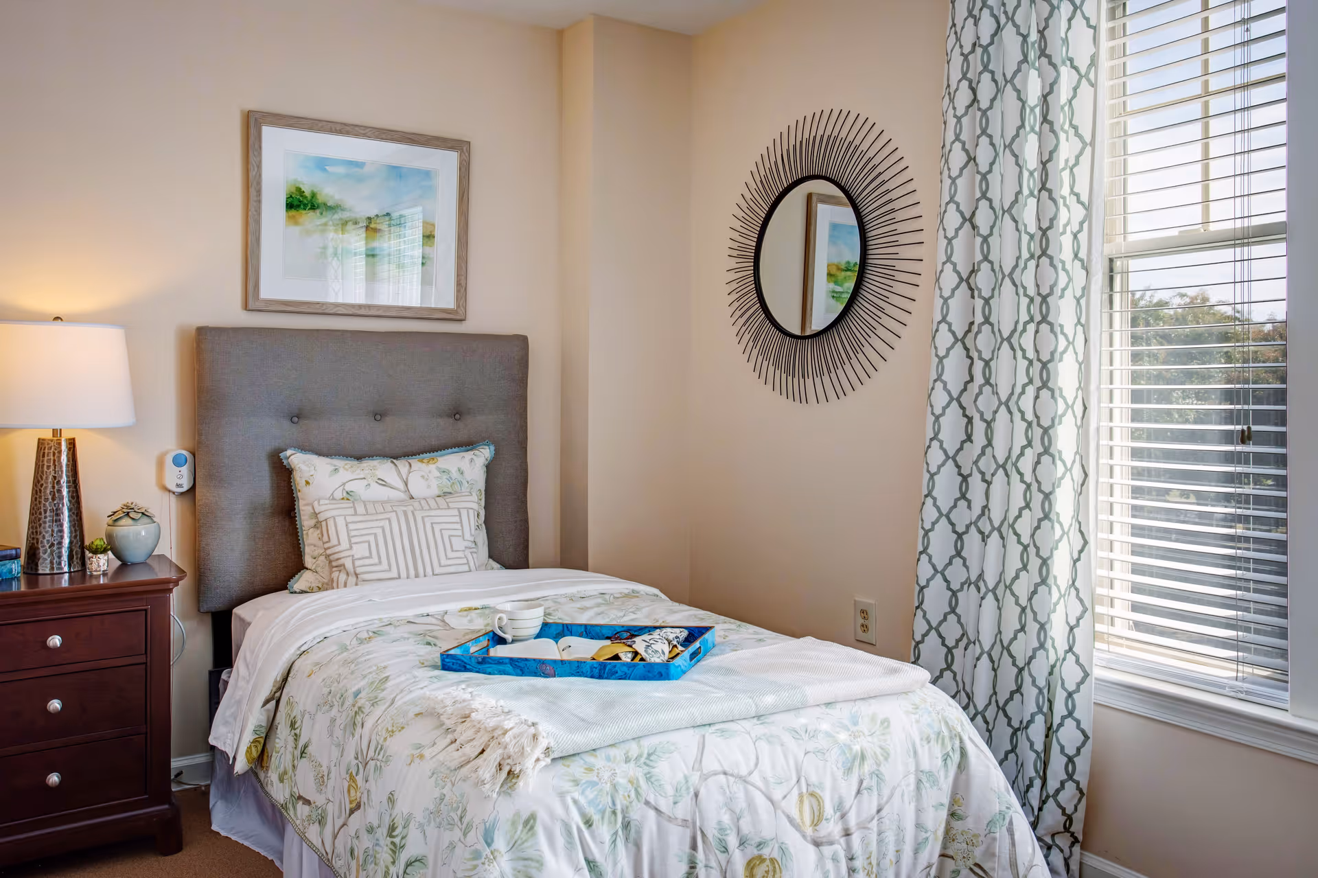A cozy bedroom with a single bed featuring a floral patterned comforter and multiple pillows. A blue tray with a cup and snacks is placed on the bed. Next to the bed is a wooden nightstand with a lamp, a small plant, and a clock. The room has a window with patterned curtains and blinds, and a decorative round mirror on the wall.
