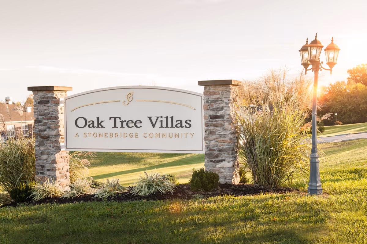 Entrance sign reading 'Oak Tree Villas A Stonebridge Community' flanked by stone pillars and a lamp post on a grassy lawn at sunset.