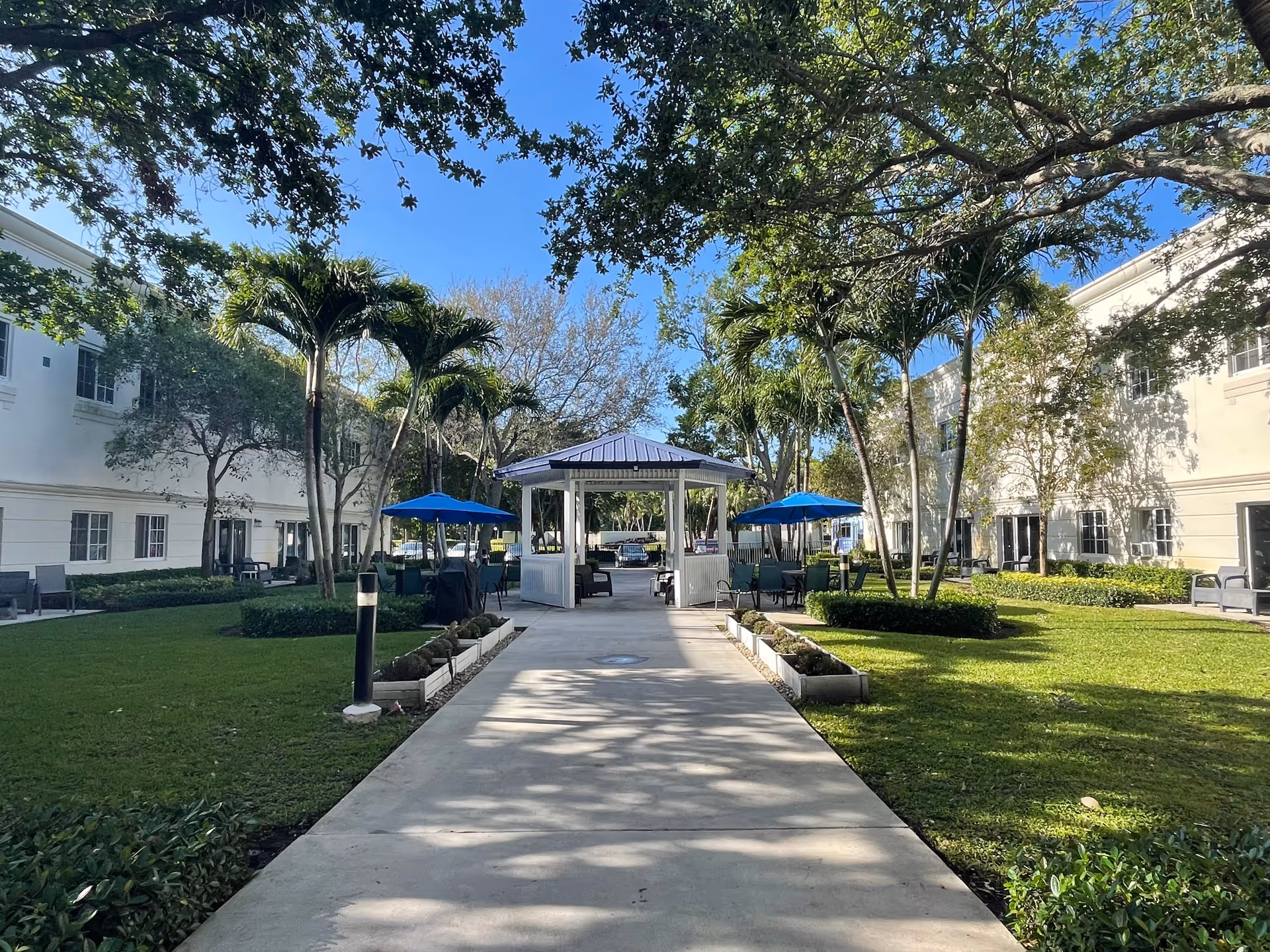 Outdoor courtyard area at Banyan Place with a paved walkway leading to a white gazebo. The courtyard is surrounded by two-story buildings with windows, green grass, palm trees, and other trees providing shade. Blue umbrellas cover seating areas near the gazebo.